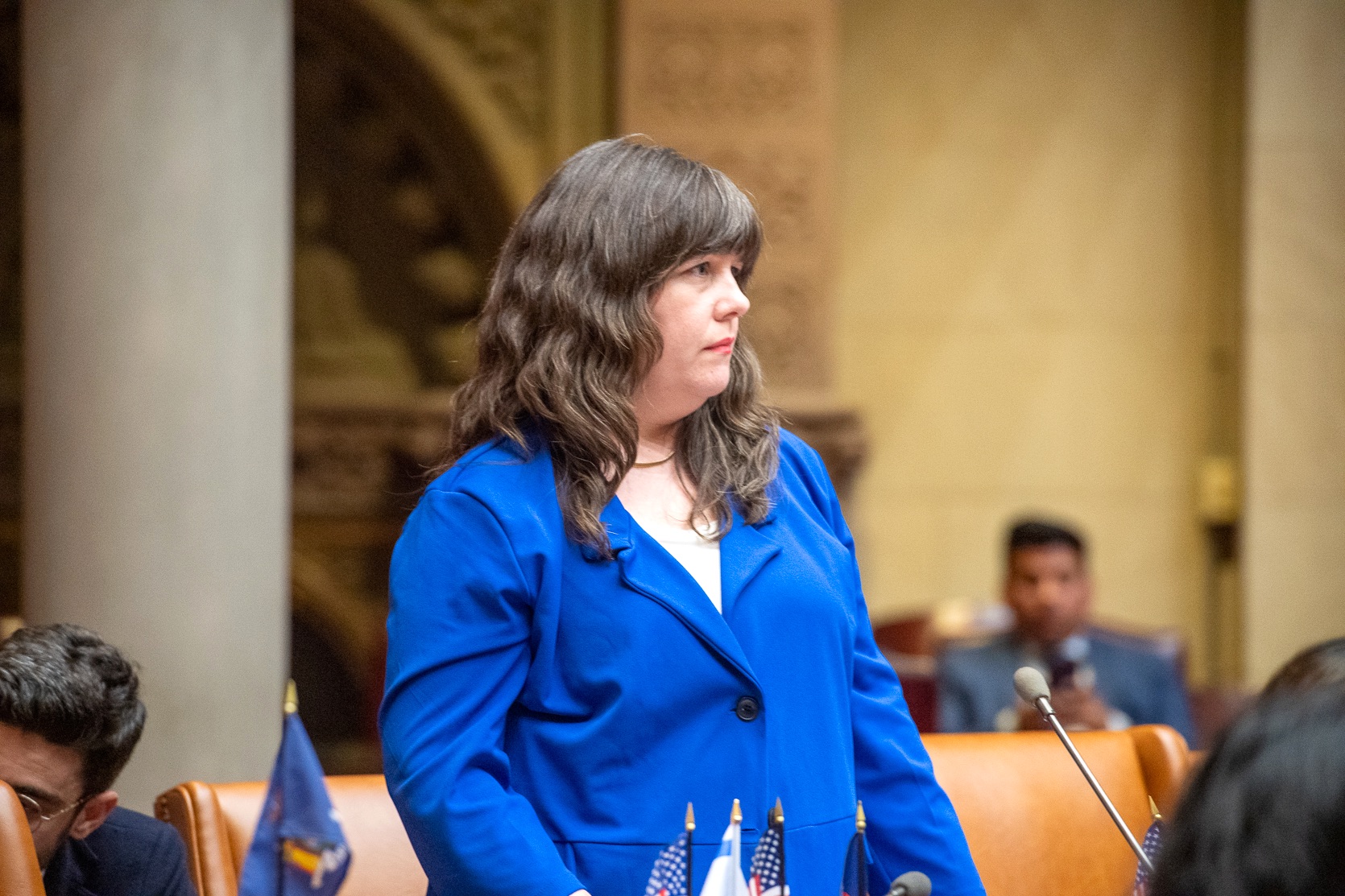 Assemblymember Gallagher stands in a neutral-toned room, wearing a blue blazer over a white shirt.