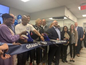 Mayor Eric Adams stands in front of a crowd of around 10 people, holding a pair of comically large scissors behind a large ribbon. The ribbon is navy blue and has white text reading "GRAND OPENING" on it twice.