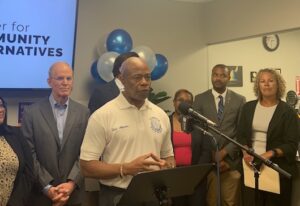 Mayor Eric Adams stands before eight other politicians and advocates, many of whom are wearing suits. He wears a white polo shirt and stands behind a small podium with a microphone attached. A television screen behind the group reads "Center for Community Alternatives."