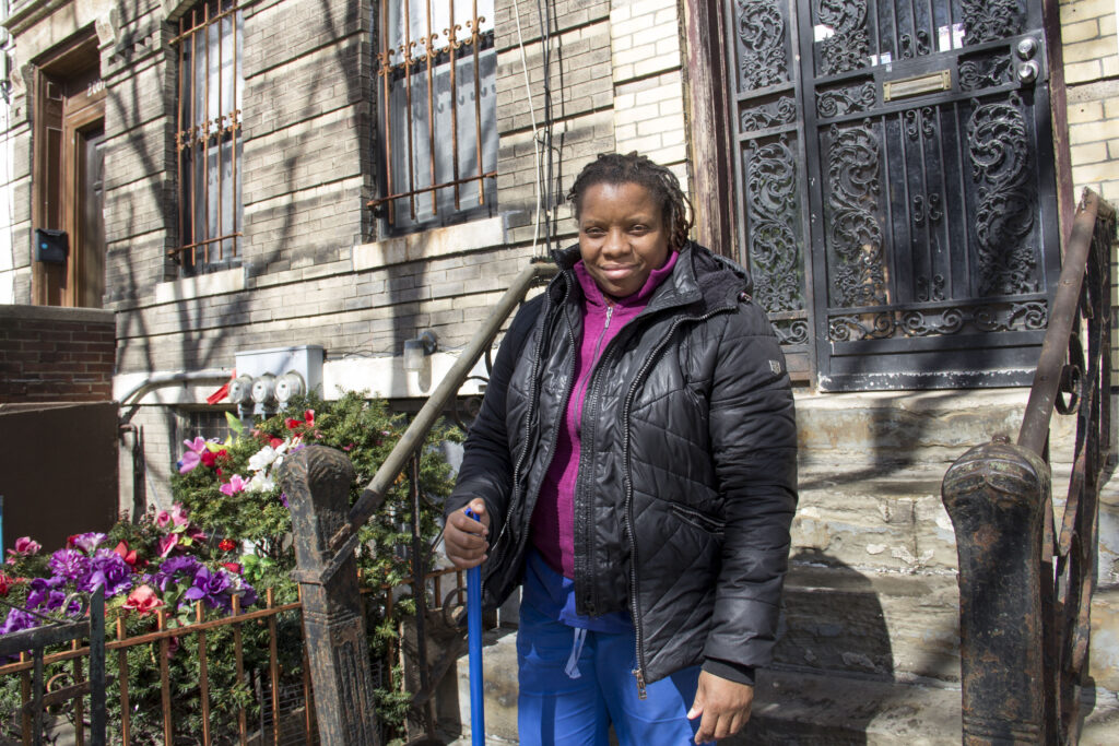 A Black woman, wearing a puffy coat and holding a rake, stands in front of an apartment.